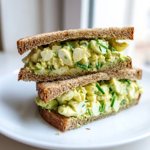 A close-up view of a creamy avocado egg salad sandwich on toasted whole grain bread, layered with fresh spinach and ripe tomato slices.